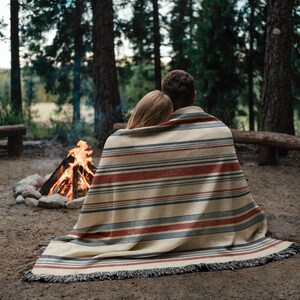 May include: A woven throw blanket featuring a horizontal striped pattern in shades of cream, muted red, and dark gray. The blanket has a textured surface and a fringed edge, draped over two individuals sitting outdoors near a campfire.
