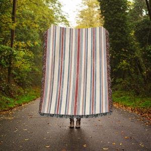 May include: A rectangular woven throw blanket featuring a vertical striped pattern in shades of cream, deep red, and navy blue. The blanket has a textured surface and frayed fringe edges along the top and bottom, with a pair of brown boots visible below.