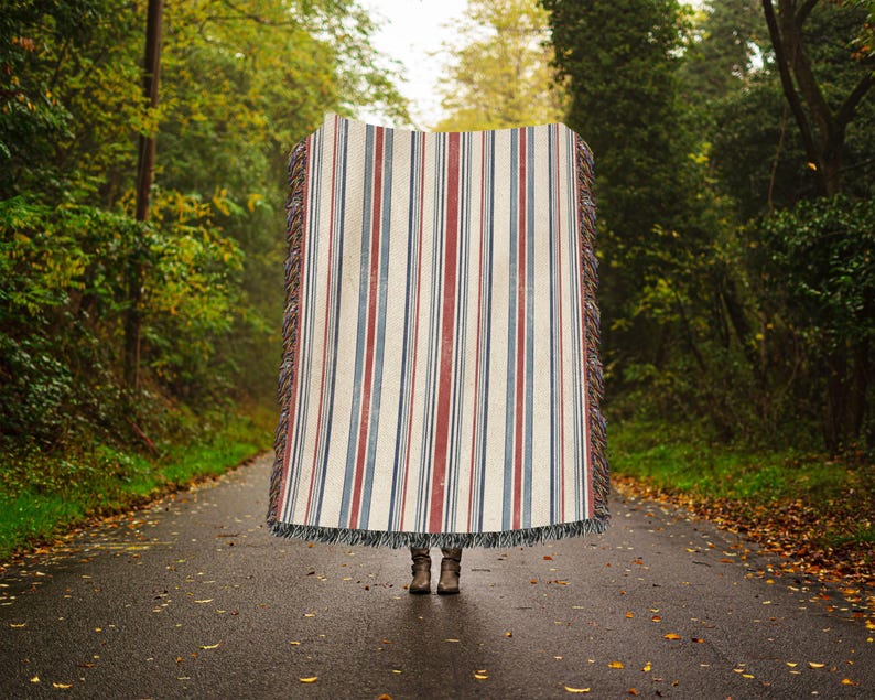 May include: A rectangular woven throw blanket featuring a vertical striped pattern in shades of cream, deep red, and navy blue. The blanket has a textured surface and frayed fringe edges along the top and bottom, with a pair of brown boots visible below.