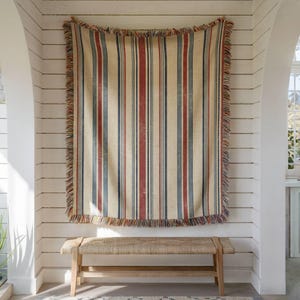 May include: A rectangular woven throw blanket featuring a vertical striped pattern in shades of cream, deep red, and slate blue. The blanket has a frayed, multi-colored fringe along all four edges and is displayed hanging vertically.