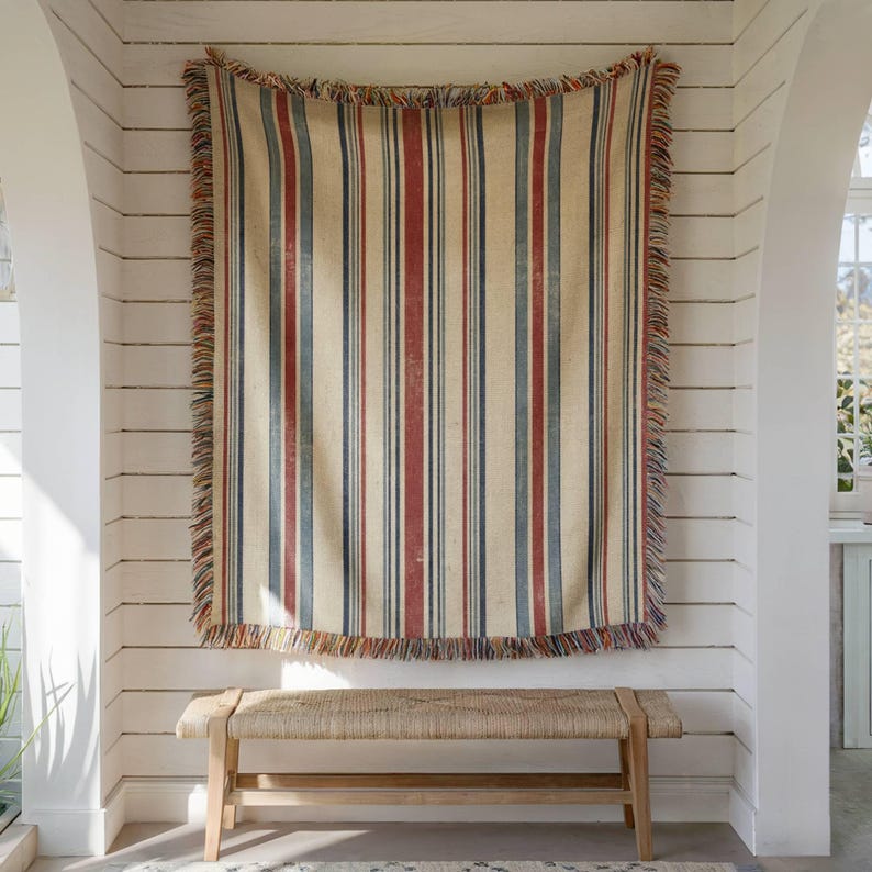 May include: A rectangular woven throw blanket featuring a vertical striped pattern in shades of cream, deep red, and slate blue. The blanket has a frayed, multi-colored fringe along all four edges and is displayed hanging vertically.