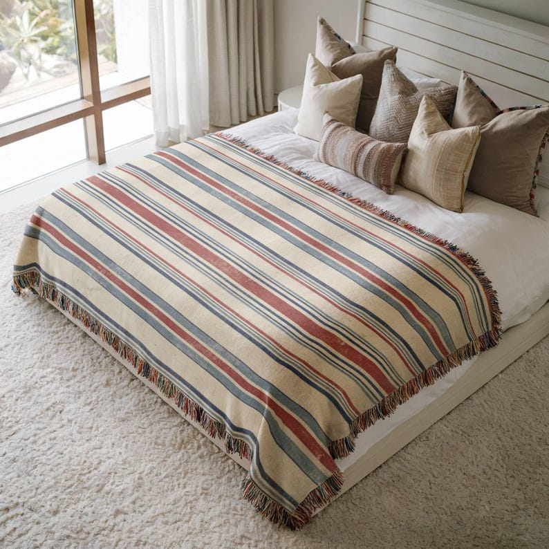 May include: A woven throw blanket featuring a pattern of horizontal stripes in shades of cream, terracotta red, and slate blue, finished with a fringed edge. The blanket is draped over a bed with white linens and several decorative throw pillows.