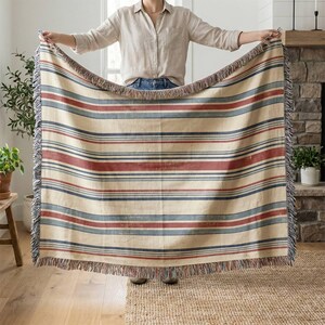 May include: A woven throw blanket featuring a horizontal striped pattern in shades of cream, muted red, and slate blue. The rectangular textile has a decorative fringe along the top and bottom edges, with a consistent striped design throughout.