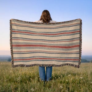 May include: A rectangular woven throw blanket featuring a repeating pattern of horizontal stripes in muted red, dark blue, and cream colors. The blanket has a textured fabric finish and is finished with a decorative fringe along the vertical edges.