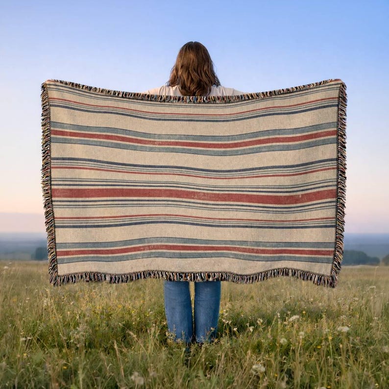 May include: A rectangular woven throw blanket featuring a repeating pattern of horizontal stripes in muted red, dark blue, and cream colors. The blanket has a textured fabric finish and is finished with a decorative fringe along the vertical edges.