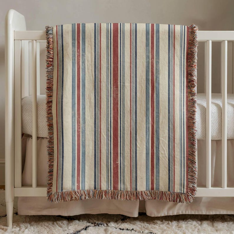 Vintage striped baby blanket with red, blue, and cream stripes draped over a white crib, perfect for Americana nursery decor and classic baby room styling.