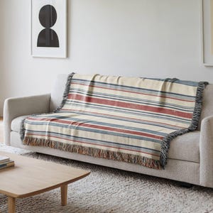 May include: A woven throw blanket featuring a horizontal striped pattern in shades of cream, muted red, and slate blue. The blanket has a textured appearance with frayed fringe edges along the bottom, draped over a light gray upholstered sofa.
