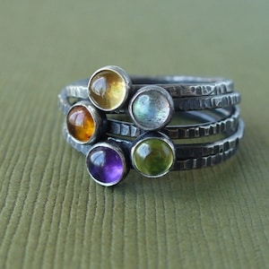 May include: A silver ring with five different colored gemstones. The gemstones are set in a hammered silver band. The ring is stacked with four other rings, creating a layered look.