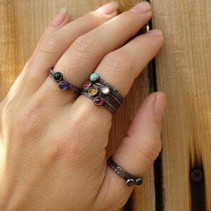 May include: A hand wearing a stack of five silver rings with colorful gemstones. The rings are on the middle finger and the thumb. The gemstones are black, turquoise, pink, yellow, and red.