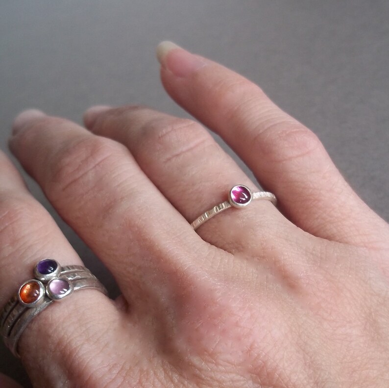 May include: Two silver rings with colourful gemstones. The top ring has a single pink gemstone. The bottom ring has three gemstones: orange, pink, and purple.