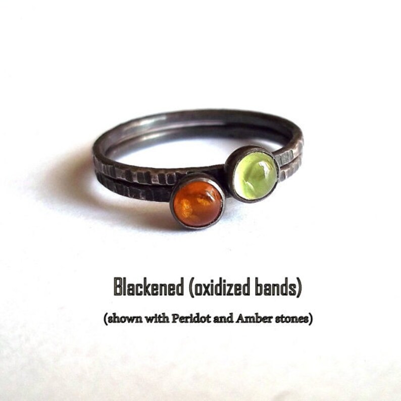 May include: Two thin black oxidised bands with a hammered texture, stacked on top of each other. The bands are adorned with a green peridot and an amber stone.