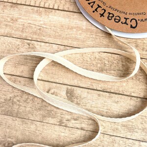 May include: A spool of natural cotton twill tape, also known as cotton webbing, on a wooden surface. The tape is a light beige color and has a woven texture. The spool is labeled "Creative!" with the website address "www.Creative!".
