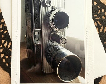 May include: A vintage silver and black film camera with a lens and a microphone. The camera is on a wooden surface.