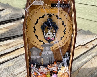 May include: A wooden coffin-shaped Halloween shadow box with a spooky theme. It features miniature figures, pumpkins, candy, and the words "Trick or Treat" and "Halloween". The interior is decorated with a doily, spiderwebs, and a skeleton.