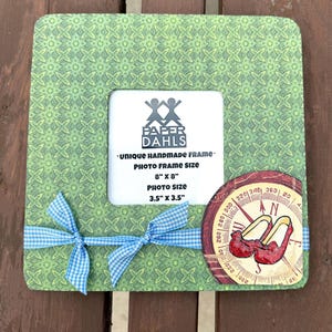 May include: A green patterned handmade photo frame with a blue gingham ribbon. The frame has a white center and a decorative compass with red shoes. The frame size is 8" x 8" and the photo size is 3.5" x 3.5". The text "PAPER DAHLS" is visible.