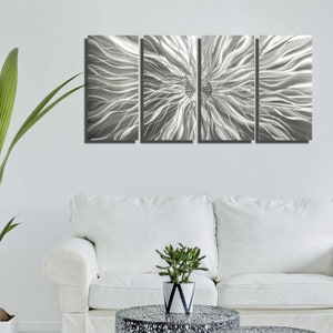 May include: Four-panel metal wall art with a silver abstract design. The artwork shows flowing, wave-like patterns radiating from a central point. The panels are mounted on a white wall above a white sofa and a small table with a plant.