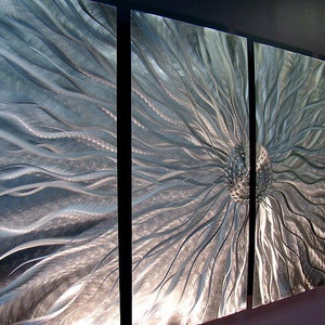 May include: Abstract metal wall art in four panels. The artwork has a central, textured design like a flower, with radiating, flowing lines in silver and grey. The panels are mounted with black spacing, creating a modern aesthetic.