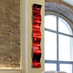 May include: A tall, wavy, red metal wall sculpture. The artwork has a glossy finish and a flowing, organic shape. The sculpture is mounted on a neutral-colored wall, with a brick wall and a large window in the background. The piece is a modern art decor.