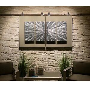 May include: Contemporary metal wall art featuring a silver floral design, split across four panels. The artwork is mounted on a neutral-toned rectangular frame, set against a textured stone wall. The piece is illuminated by soft lighting, creating a modern aesthetic.