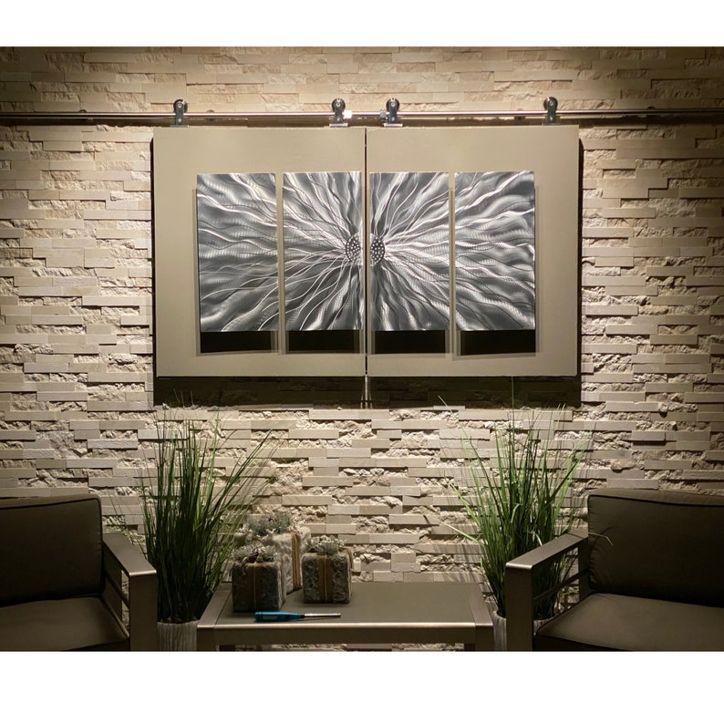 May include: Contemporary metal wall art featuring a silver floral design, split across four panels. The artwork is mounted on a neutral-toned rectangular frame, set against a textured stone wall. The piece is illuminated by soft lighting, creating a modern aesthetic.