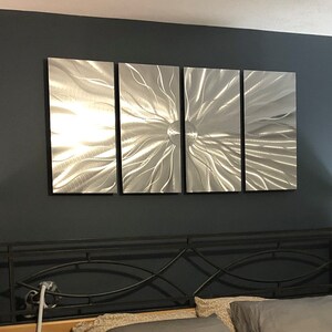 May include: A bedroom showcases a four-piece metal wall art installation with a textured, radial pattern. The artwork is mounted on a dark grey wall above a bed with a black metal headboard and wooden posts. Two lamps with patterned shades are on either side.