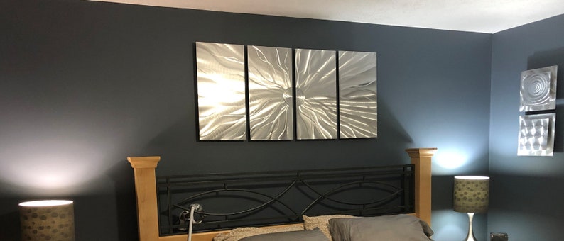 May include: A bedroom showcases a four-piece metal wall art installation with a textured, radial pattern. The artwork is mounted on a dark grey wall above a bed with a black metal headboard and wooden posts. Two lamps with patterned shades are on either side.