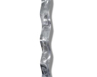 May include: A brushed metal wall sculpture with a wavy, vertical design. The silver artwork features circular and linear patterns, creating a textured appearance. The piece is mounted on a white background, casting a shadow.
