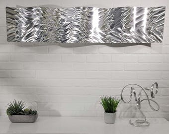 Puede incluir: Una gran escultura de pared abstracta de metal con un diseño ondulado. La escultura es plateada y cuelga de una pared de ladrillo blanco. Debajo de la escultura hay un mueble blanco con una jardinera de suculentas, una pequeña planta en maceta y una escultura decorativa de metal.