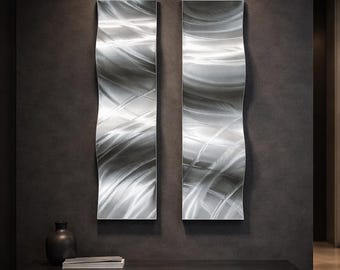 May include: Two abstract metal wall art pieces with a wavy design. The artwork has a brushed silver finish, creating a dynamic interplay of light and shadow. The pieces are mounted on a dark grey wall, adding a modern touch to the space.