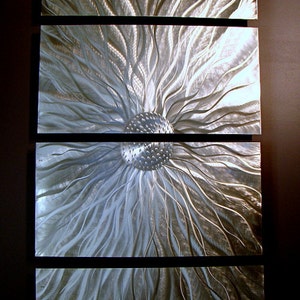 May include: Abstract metal wall art comprising four rectangular panels. Each panel displays a textured, flowing design in silver and grey tones, reminiscent of stylized floral or sunburst patterns. The artwork is mounted on a dark background.