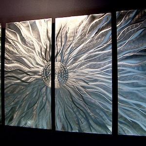 May include: Abstract metal wall art in four panels. The artwork shows a central, textured sphere with radiating lines and a metallic silver and blue colour palette. The panels are mounted on a dark wall.