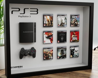 May include: A framed display featuring a black PlayStation 3 console, controller, and game cases. The games include "The Last of Us," "Assassin's Creed," and "Far Cry 3." The frame is black, and the background is white.