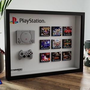 May include: A framed display of a grey PlayStation console, a grey controller, and nine game cartridges. The frame is black and the background is white. The text "PlayStation" is printed in black on the white background.
