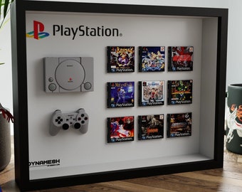May include: A framed PlayStation display featuring a gray console, controller, and game cases. The PlayStation logo is visible at the top. The frame is black, and the background is white. A Spider-Man figure and a mug are also in the image.