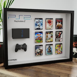Peut inclure: Une vitrine noire encadrée avec une console PlayStation 2, une manette et neuf cartouches de jeux vidéo. La vitrine porte l'inscription "PS2 PlayStation 2" en haut.