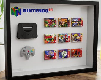 May include: A framed display featuring a black Nintendo 64 console, a gray controller, and several game cartridges. The Nintendo 64 logo is visible. The display is set against a white background within a black frame.