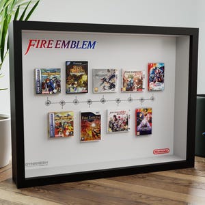 Peut inclure: Présentation encadrée noire avec neuf boîtiers de jeux vidéo Fire Emblem, disposés chronologiquement. Le logo Fire Emblem est en haut. Un mug noir avec une illustration de personnage est à droite, près d'une plante en pot.