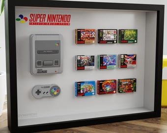 May include: A framed Super Nintendo display featuring a gray console, controller, and game cartridges. The Super Nintendo logo is visible at the top. The display is in a black frame, with a yellow question mark block in the background.