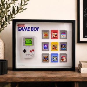 Op de afbeelding: Een zwarte schaduwdooslijst met een lichtgrijze Nintendo Game Boy handheld console en negen kleurrijke spelcartridges in een raster. Het Game Boy-scherm toont tekst en het Nintendo Game Boy-logo is in blauw en rood afgedrukt.