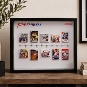 Peut inclure: Un cadre mural noir présentant un fond blanc avec le logo Fire Emblem et la marque Nintendo. Dix couvertures de jeux miniatures sont disposées sur deux rangées le long d'un graphique chronologique, illustrant divers titres de la série.