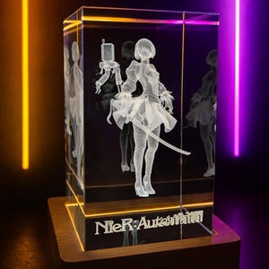 Peut inclure: Un bloc de cristal rectangulaire transparent avec une image gravée en 3D d'un personnage d'anime féminin tenant une épée. La base est en bois et le texte "NieR:Automata" est visible. Le bloc est éclairé par des néons orange et violets.