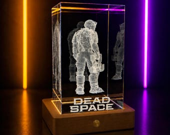 May include: A clear, rectangular glass block with a 3D etched image of a figure in a spacesuit. The block sits on a wooden base with the words "DEAD SPACE" etched on the front. The background is dark with orange and purple neon lights.