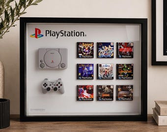 May include: A black framed shadow box display featuring a miniature grey PlayStation console and controller, accompanied by nine small game case replicas arranged in a grid, all set against a white background with the PlayStation logo at the top.