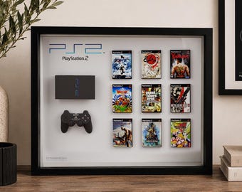 May include: A black shadow box frame containing a PlayStation 2 console, a black controller, and nine miniature game cases arranged in a grid. The white background features the PS2 and PlayStation 2 logos in blue and black text respectively.
