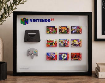 May include: A black shadow box frame containing a Nintendo 64 logo, a black console shell, a gray controller, and nine miniature game cartridges arranged in a grid. The display features colorful game labels and the text NINTENDO 64 at the top.