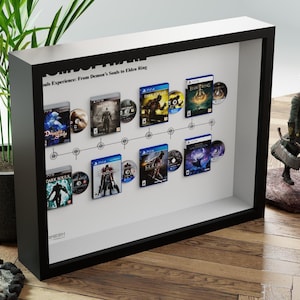 May include: A framed display featuring video game cases and discs from the "Souls" series, including "Demon's Souls" and "Elden Ring." The black frame houses the games, arranged along a timeline. A figurine stands to the right.