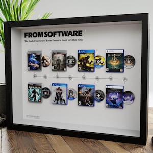 May include: Framed artwork featuring video game covers and discs from the "Souls" series, including "Dark Souls" and "Elden Ring." The black frame displays the games in chronological order, with the text "FROM SOFTWARE" at the top.