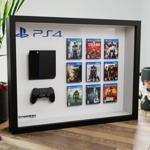 Peut inclure: Un cadre de présentation noir de type boîte vitrine contenant une console, une manette et neuf boîtiers de jeux fixés sur un fond blanc.