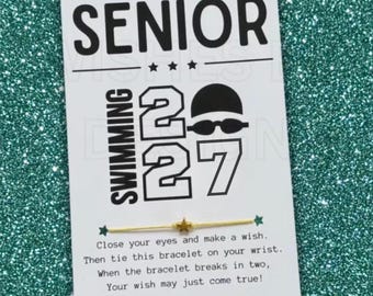 May include: A thin gold-colored string bracelet with a small gold star charm, attached to a white card featuring the text SENIOR SWIMMING 27, a graphic of a swimmer, and instructions to close your eyes, make a wish, and tie the bracelet on your wrist.