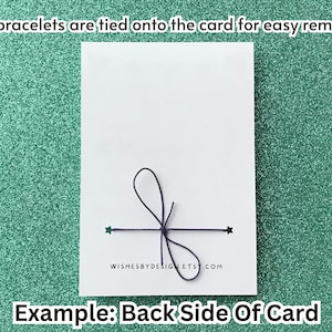 Peut inclure: Une carte blanche avec un n&oelig;ud noir et des d&eacute;tails de ficelle, avec le texte "WISHESBYDESIGNETS.COM". Le texte "Les bracelets sont attach&eacute;s &agrave; la carte pour un retrait facile !" est en haut, et "Exemple : Verso de la carte" est en bas. Le fond est un vert d'eau scintillant.